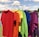 A row of colorful t-shirts hanging on a clothesline outdoors, with a scenic background of a lake and trees under a partly cloudy sky. The shirts are arranged in a rainbow sequence, showcasing red, orange, yellow, green, blue, and purple hues.