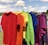 A row of colorful t-shirts hanging on a clothesline outdoors, with a scenic background of a lake and trees under a partly cloudy sky. The shirts are arranged in a rainbow sequence, showcasing red, orange, yellow, green, blue, and purple hues.