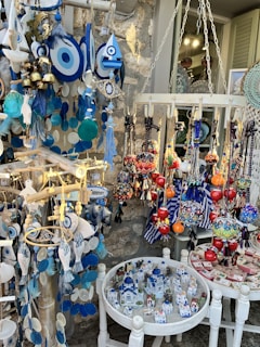 A colorful display of souvenirs and decorative items, featuring Evil Eye ornaments in blue and white, vibrant hanging glass spheres, and miniature models of traditional white and blue houses resembling those from the Mediterranean. The collection includes various handcrafted items like bells, dream catchers, and fish-shaped decorations, all arranged against a rustic stone wall.