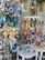 A colorful display of souvenirs and decorative items, featuring Evil Eye ornaments in blue and white, vibrant hanging glass spheres, and miniature models of traditional white and blue houses resembling those from the Mediterranean. The collection includes various handcrafted items like bells, dream catchers, and fish-shaped decorations, all arranged against a rustic stone wall.