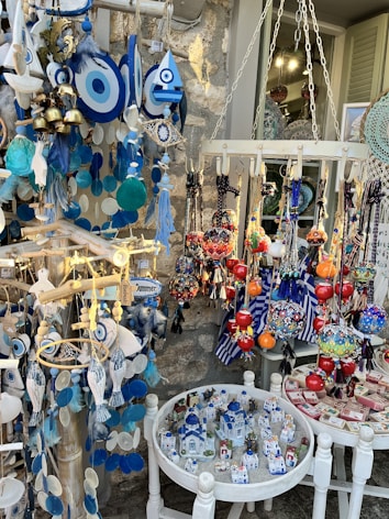 A colorful display of souvenirs and decorative items, featuring Evil Eye ornaments in blue and white, vibrant hanging glass spheres, and miniature models of traditional white and blue houses resembling those from the Mediterranean. The collection includes various handcrafted items like bells, dream catchers, and fish-shaped decorations, all arranged against a rustic stone wall.
