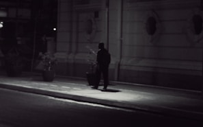 A shadowy figure tailing a person along a narrow European street during twilight.