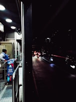 A dimly lit urban scene with a security guard standing near a booth on a pedestrian walkway, checking their phone. Beyond the booth, several motorcycles with their headlights on are visible on a wet road, creating reflections. The area is partially illuminated by overhead lights.