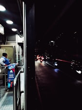 A dimly lit urban scene with a security guard standing near a booth on a pedestrian walkway, checking their phone. Beyond the booth, several motorcycles with their headlights on are visible on a wet road, creating reflections. The area is partially illuminated by overhead lights.