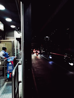 A dimly lit urban scene with a security guard standing near a booth on a pedestrian walkway, checking their phone. Beyond the booth, several motorcycles with their headlights on are visible on a wet road, creating reflections. The area is partially illuminated by overhead lights.