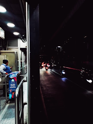 A dimly lit urban scene with a security guard standing near a booth on a pedestrian walkway, checking their phone. Beyond the booth, several motorcycles with their headlights on are visible on a wet road, creating reflections. The area is partially illuminated by overhead lights.