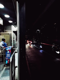A dimly lit urban scene with a security guard standing near a booth on a pedestrian walkway, checking their phone. Beyond the booth, several motorcycles with their headlights on are visible on a wet road, creating reflections. The area is partially illuminated by overhead lights.