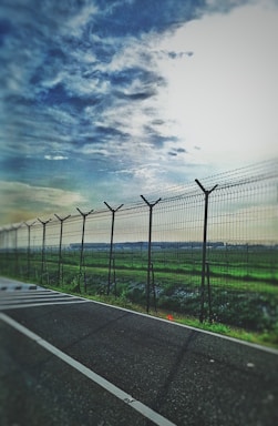 A wire fence stretches alongside a road, separating it from a vast field. The sky above is partly cloudy with blue and white hues, creating a calm atmosphere.