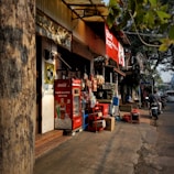 A small street-side shop with a variety of items displayed. The shopfront features a red Coca-Cola refrigerator and a stack of boxes. Various snacks hang for sale, and there are advertisements visible. A tree partially obscures the view, and a motorcycle is parked on the street nearby.