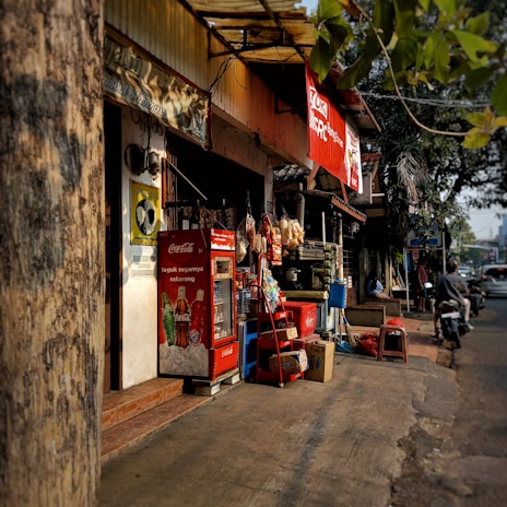 A small street-side shop with a variety of items displayed. The shopfront features a red Coca-Cola refrigerator and a stack of boxes. Various snacks hang for sale, and there are advertisements visible. A tree partially obscures the view, and a motorcycle is parked on the street nearby.