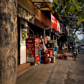 A small street-side shop with a variety of items displayed. The shopfront features a red Coca-Cola refrigerator and a stack of boxes. Various snacks hang for sale, and there are advertisements visible. A tree partially obscures the view, and a motorcycle is parked on the street nearby.