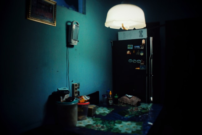 A dimly lit kitchen with a dark teal wall partially illuminated by a hanging ceiling lamp. On the left, a wall-mounted appliance, possibly a phone or intercom, hangs above a counter cluttered with various kitchen items, including containers and a cutting board. The right side features a black refrigerator adorned with magnets and notes.