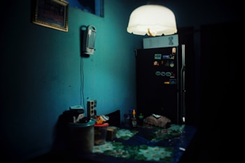 A dimly lit kitchen with a dark teal wall partially illuminated by a hanging ceiling lamp. On the left, a wall-mounted appliance, possibly a phone or intercom, hangs above a counter cluttered with various kitchen items, including containers and a cutting board. The right side features a black refrigerator adorned with magnets and notes.