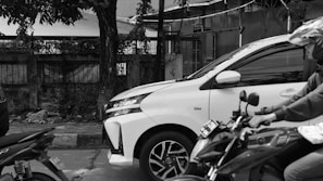 A busy urban street setting with a motorcycle stopped alongside a white car. A tree is prominently positioned next to a fence, casting shadows across the sidewalk. Motorcyclists wear helmets, hinting at a sense of safety and traffic regulation.