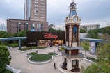 A bustling plaza features a prominent clock tower with intricate design details, surrounded by greenery and pathways. In the background, a large building with the Disney logo at the entrance attracts visitors. People are casually walking and gathering in the open space, enjoying a relaxed atmosphere.