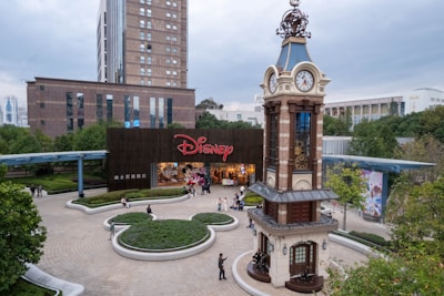 A bustling plaza features a prominent clock tower with intricate design details, surrounded by greenery and pathways. In the background, a large building with the Disney logo at the entrance attracts visitors. People are casually walking and gathering in the open space, enjoying a relaxed atmosphere.