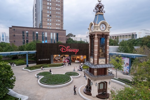 A bustling plaza features a prominent clock tower with intricate design details, surrounded by greenery and pathways. In the background, a large building with the Disney logo at the entrance attracts visitors. People are casually walking and gathering in the open space, enjoying a relaxed atmosphere.