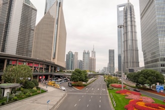 A modern cityscape featuring tall skyscrapers, including distinctive architectural designs. The foreground includes a well-maintained road with several lanes and colorful landscaped flower beds alongside it. There are also visible vehicles, buses, and pedestrians near a bus stop, adding urban vibrancy.