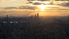 The image captures a city skyline at sunset, with tall skyscrapers silhouetted against a golden sky. The sun is setting on the horizon, casting a warm glow and illuminating scattered clouds. The urban landscape is expansive, with a river and bridges visible in the distance.