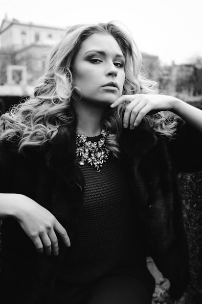 A woman with wavy hair poses confidently in a cozy, fur-like coat and a detailed necklace. The black and white tone adds a classic and dramatic effect to the image, with a blurred architectural background that suggests an outdoor setting.