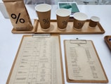A selection of coffee items on a white countertop includes a bag of coffee, four paper cups with a percentage symbol, two clipboards with menus or price lists, tablets in the background likely for orders, and a hint of electronic equipment for transactions.