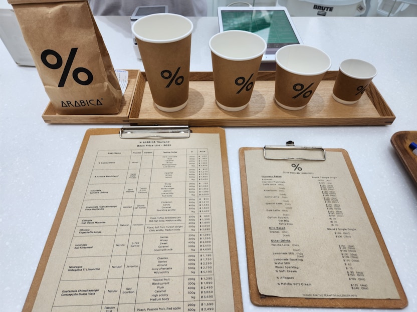 A selection of coffee items on a white countertop includes a bag of coffee, four paper cups with a percentage symbol, two clipboards with menus or price lists, tablets in the background likely for orders, and a hint of electronic equipment for transactions.