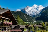 A picturesque alpine village is nestled in a valley with wooden chalet-style houses adorned with flower boxes. Snow-capped mountains tower majestically in the background under a clear blue sky.