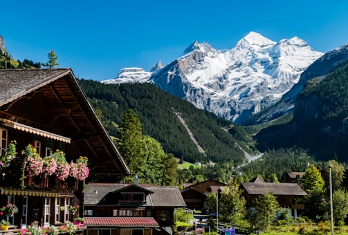 A picturesque alpine village is nestled in a valley with wooden chalet-style houses adorned with flower boxes. Snow-capped mountains tower majestically in the background under a clear blue sky.