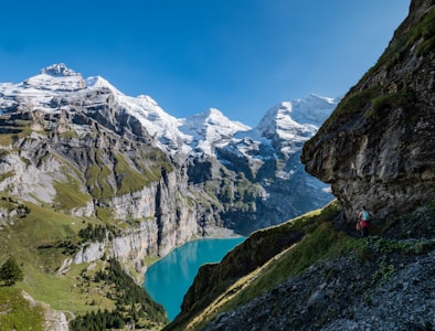 A breathtaking mountainous landscape with towering snow-capped peaks, lush green valleys, and a crystal-clear blue lake nestled in the valley. A hiker with a backpack is seen on a path along a rocky incline, adding a sense of scale to the vastness of the natural scenery.