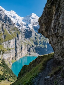 A stunning alpine landscape featuring snow-capped mountains towering above a turquoise-blue lake. The rugged cliffs are adorned with patches of green vegetation, and a narrow, rocky trail winds along the side of the hill. The sky above is clear and bright blue, adding to the serene and majestic atmosphere.