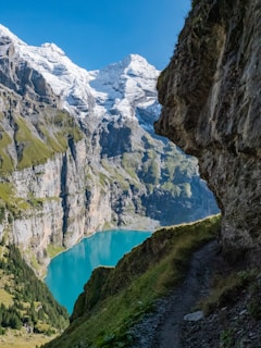 A stunning alpine landscape featuring snow-capped mountains towering above a turquoise-blue lake. The rugged cliffs are adorned with patches of green vegetation, and a narrow, rocky trail winds along the side of the hill. The sky above is clear and bright blue, adding to the serene and majestic atmosphere.
