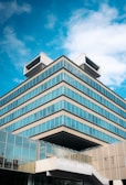A modern multi-story building with a unique architectural design. The structure features stacked, rectangular layers with extensive use of glass windows, allowing for a reflective aesthetic. The sky in the background is clear with scattered clouds, providing a backdrop of bright blue.