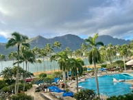 A tropical resort setting featuring a large swimming pool surrounded by lounge chairs and umbrellas. Tall palm trees are scattered throughout the area, with lush greenery and colorful flowers enhancing the scenery. In the background, there are majestic mountains partially covered by clouds, and a calm body of water is visible, complementing the serene atmosphere.