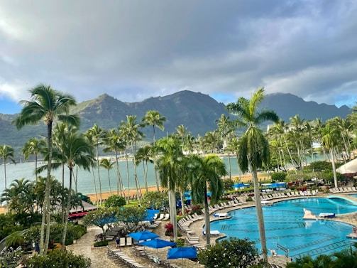 A tropical resort setting featuring a large swimming pool surrounded by lounge chairs and umbrellas. Tall palm trees are scattered throughout the area, with lush greenery and colorful flowers enhancing the scenery. In the background, there are majestic mountains partially covered by clouds, and a calm body of water is visible, complementing the serene atmosphere.