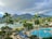 A tropical resort setting featuring a large swimming pool surrounded by lounge chairs and umbrellas. Tall palm trees are scattered throughout the area, with lush greenery and colorful flowers enhancing the scenery. In the background, there are majestic mountains partially covered by clouds, and a calm body of water is visible, complementing the serene atmosphere.
