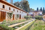 A beautifully landscaped garden features a long pathway flanked by green hedges and lined with water fountains. The area is surrounded by stone buildings with wooden shutters and climbing plants. A backdrop of tall trees and bright blue skies enhances the tranquil atmosphere.