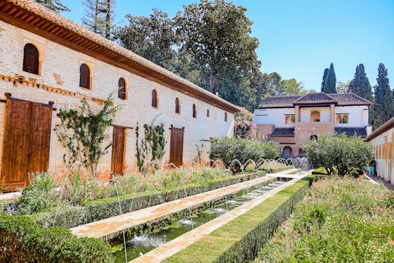 A beautifully landscaped garden features a long pathway flanked by green hedges and lined with water fountains. The area is surrounded by stone buildings with wooden shutters and climbing plants. A backdrop of tall trees and bright blue skies enhances the tranquil atmosphere.