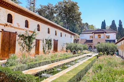 A beautifully landscaped garden features a long pathway flanked by green hedges and lined with water fountains. The area is surrounded by stone buildings with wooden shutters and climbing plants. A backdrop of tall trees and bright blue skies enhances the tranquil atmosphere.