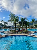 A luxurious outdoor swimming pool area with clear blue water, surrounded by tropical palm trees and vibrant flowering plants. Lounge chairs are arranged around the pool, providing a relaxing space for sunbathing. The scene is set against a backdrop of green hills and a partly cloudy sky with patches of blue.