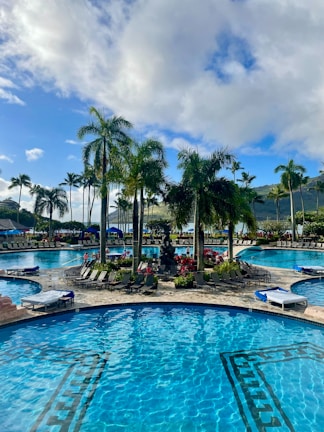 A luxurious outdoor swimming pool area with clear blue water, surrounded by tropical palm trees and vibrant flowering plants. Lounge chairs are arranged around the pool, providing a relaxing space for sunbathing. The scene is set against a backdrop of green hills and a partly cloudy sky with patches of blue.