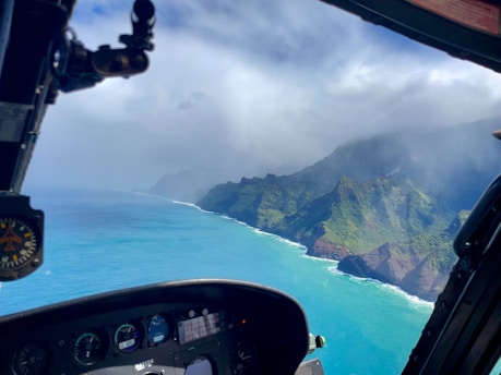 An aerial view from a helicopter cockpit overlooking a breathtaking coastal landscape. The expansive blue ocean meets dramatic, green-covered cliffs under a partly cloudy sky. The helicopter's controls and instruments are visible in the foreground, giving a sense of altitude and perspective.