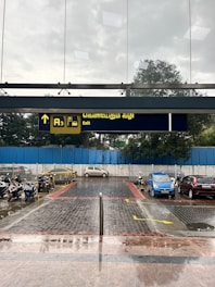 A paved area leads to a parking lot with motorcycles and parked cars. Above, a sign with text in Tamil and English indicates an exit. The sky is overcast, suggesting possible rain, and there is a blue fence in the background.
