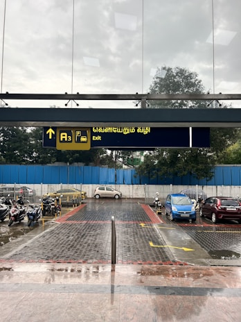 A paved area leads to a parking lot with motorcycles and parked cars. Above, a sign with text in Tamil and English indicates an exit. The sky is overcast, suggesting possible rain, and there is a blue fence in the background.