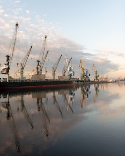 Several large industrial cranes stand along the dockside over a calm body of water. A cargo ship is moored alongside, and the cranes are poised above it, possibly for loading or unloading containers. The reflection of the cranes and the lightly clouded sky is visible in the water, creating a symmetrical visual effect.