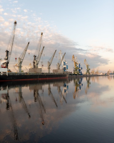 Several large industrial cranes stand along the dockside over a calm body of water. A cargo ship is moored alongside, and the cranes are poised above it, possibly for loading or unloading containers. The reflection of the cranes and the lightly clouded sky is visible in the water, creating a symmetrical visual effect.