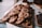 A wooden tray is filled with freshly baked cookies topped with chunks of chocolate and nuts. The cookies have a golden-brown texture and appear soft and chewy. Beside them, slices of a dark loaf cake are arranged on another wooden board. The background suggests a cozy bakery setting.