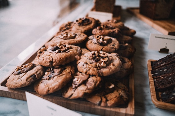 A wooden tray is filled with freshly baked cookies topped with chunks of chocolate and nuts. The cookies have a golden-brown texture and appear soft and chewy. Beside them, slices of a dark loaf cake are arranged on another wooden board. The background suggests a cozy bakery setting.