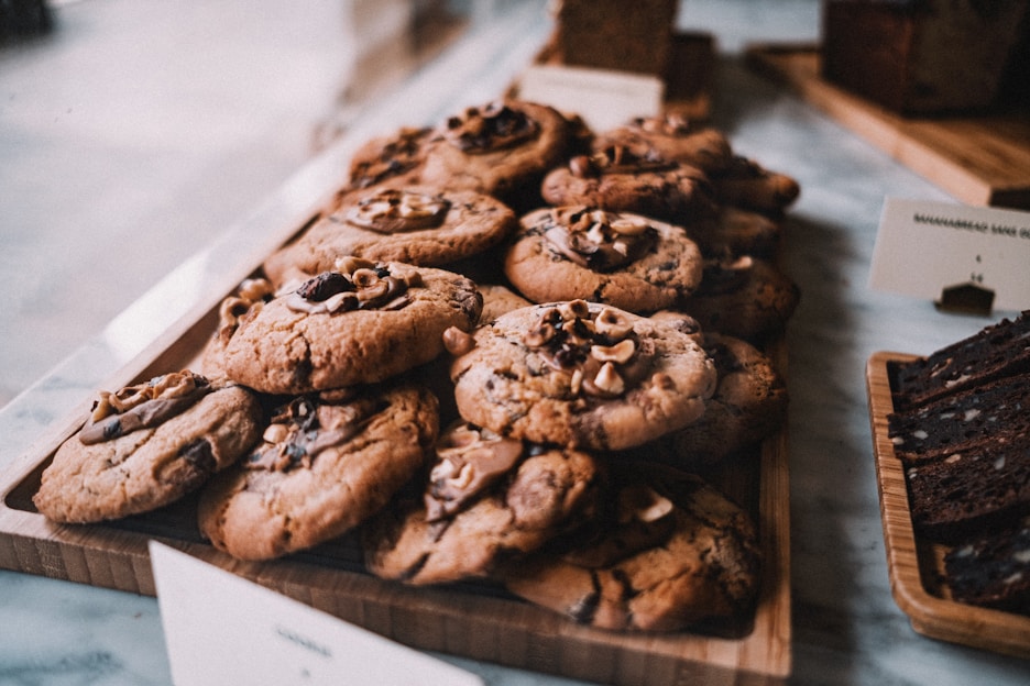 A wooden tray is filled with freshly baked cookies topped with chunks of chocolate and nuts. The cookies have a golden-brown texture and appear soft and chewy. Beside them, slices of a dark loaf cake are arranged on another wooden board. The background suggests a cozy bakery setting.