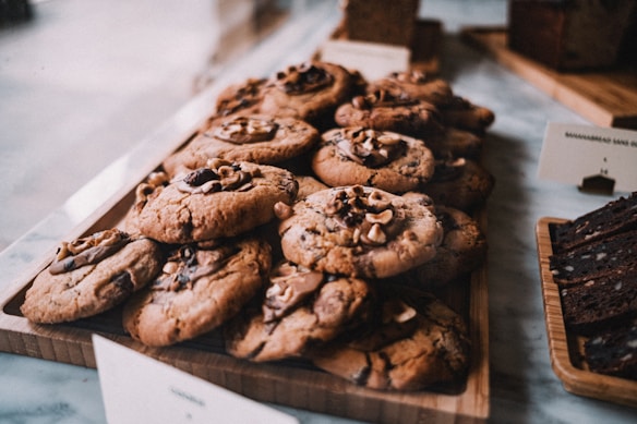 A wooden tray is filled with freshly baked cookies topped with chunks of chocolate and nuts. The cookies have a golden-brown texture and appear soft and chewy. Beside them, slices of a dark loaf cake are arranged on another wooden board. The background suggests a cozy bakery setting.