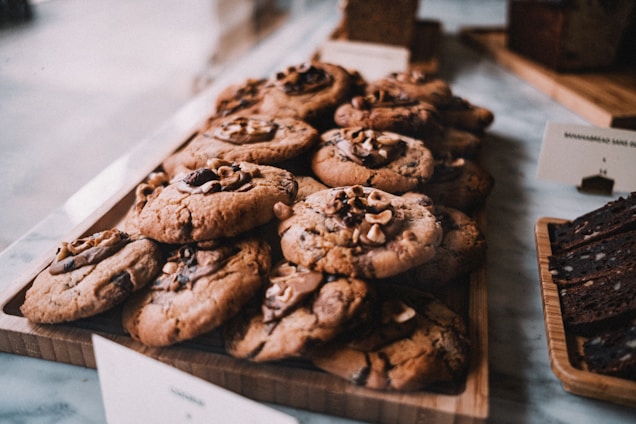 A wooden tray is filled with freshly baked cookies topped with chunks of chocolate and nuts. The cookies have a golden-brown texture and appear soft and chewy. Beside them, slices of a dark loaf cake are arranged on another wooden board. The background suggests a cozy bakery setting.